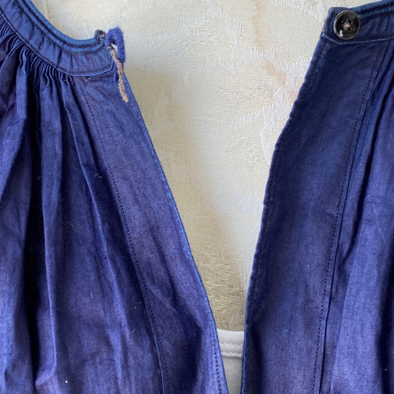 May include: Close-up of the neckline of a dark blue denim dress with a button closure and a gathered detail at the top.