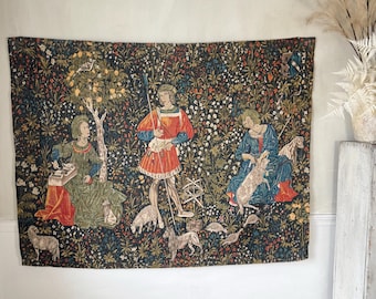 May include: A tapestry depicting a scene from the Lady and the Unicorn series. The tapestry features three figures in a forest setting, with a variety of flowers and animals. The figures are dressed in medieval clothing and are engaged in various activities. The tapestry is woven in a variety of colors, including red, blue, green, and yellow.