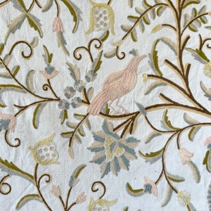 May include: A white fabric with embroidered floral and bird designs in shades of green, brown, pink, and blue.