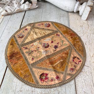 Puede incluir: Textil redondo de estilo antiguo con diseño de patchwork. El centro es de color melocotón con motivos florales bordados en rojo, verde y dorado. El borde exterior es marrón oscuro con un borde cosido. Probablemente un salvamanteles o una pieza decorativa.