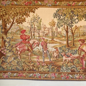 May include: A tapestry depicting a hunting scene with figures in period clothing, horses, and dogs. The artwork features a castle in the background, surrounded by trees and a floral border in shades of brown, green, and red.