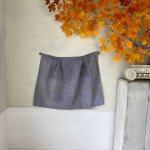 May include: A grey linen apron with a gathered waist and two pockets. The apron is hanging on a white wall.