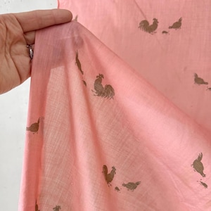 May include: Pale pink fabric featuring a repeating pattern of brown rooster and hen illustrations. The fabric is held up, revealing its texture and translucence. Ideal for crafting or home decor projects.