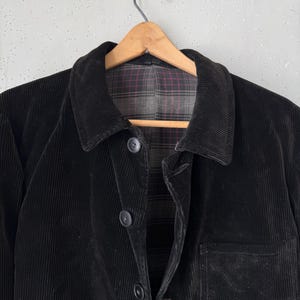 Op de afbeelding: Zwarte corduroy jas met een klassieke kraag en knoopsluiting. De jas heeft een enkele borstzak en een geruite voering zichtbaar bij de kraag. De jas hangt aan een houten kleerhanger.
