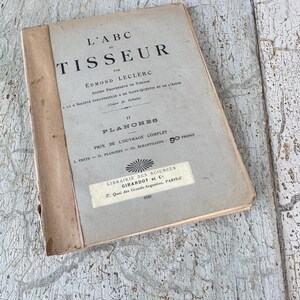 Pode incluir: Um livro vintage intitulado "L'ABC du TISSEUR" de Edmond Leclerc. O livro &eacute; um guia para tecer e apresenta ilustra&ccedil;&otilde;es. A capa &eacute; bege com texto preto e um pequeno adesivo que diz "LIBRAIRIE DES SCIENCES GIRARDOT et C 27, Quai des Grands Augustins, PARIS 1923."