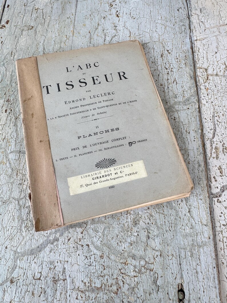 Pode incluir: Um livro vintage intitulado "L'ABC du TISSEUR" de Edmond Leclerc. O livro &eacute; um guia para tecer e apresenta ilustra&ccedil;&otilde;es. A capa &eacute; bege com texto preto e um pequeno adesivo que diz "LIBRAIRIE DES SCIENCES GIRARDOT et C 27, Quai des Grands Augustins, PARIS 1923."