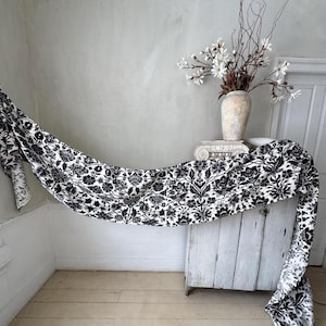 Op de afbeelding: Een zwart-wit bloemenpatroon stof gedrapeerd over een witte kast. Een vaas met witte bloemen en takken staat bovenop de kast. De stof heeft een gedetailleerd, herhalend ontwerp.
