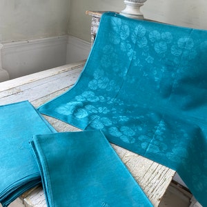 Könnte beinhalten: Ein Set tealfarbener Leinenservietten mit gestickten Initialen. Die Servietten sind gefaltet und auf einem Holztisch gestapelt. Im Hintergrund ist auch eine tealfarbene Tischdecke mit Blumenmuster zu sehen.