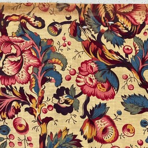 Op de afbeelding: Close-up van een stof met een herhalend bloemenpatroon. Het ontwerp toont grote, gestileerde bloemen in tinten roze, rood, blauw en geel op een crèmekleurige achtergrond. De stof heeft een vintage of antieke uitstraling.