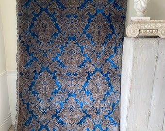 May include: A large, blue velvet curtain with a repeating brown damask pattern. The curtain has a fringed edge on the left side. A white decorative element is visible in the background.