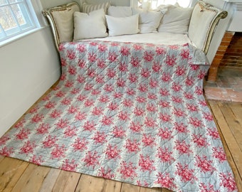 Puede incluir: Una colcha floral vintage con un patrón floral gris y rosa. La colcha está acolchada en forma de diamante y tiene un borde deshilachado. La colcha está sobre un suelo de madera.