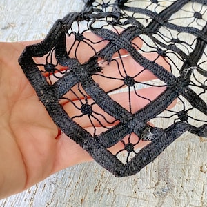 May include: Black lace trim with a geometric pattern. The trim is made of thick black cord and features a repeating design of squares and circles.