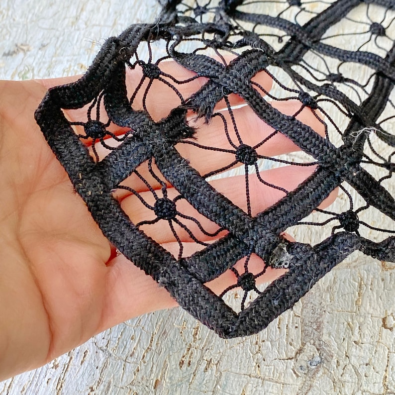 May include: Black lace trim with a geometric pattern. The trim is made of thick black cord and features a repeating design of squares and circles.