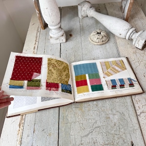 Puede incluir: Un libro abierto que muestra muestras de tela en varios colores y patrones. Las muestras incluyen rojo con lunares blancos, dorado floral y rayas azules y amarillas. El libro está sobre una superficie de madera clara y desgastada.