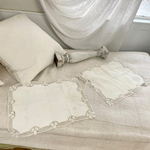 May include: Two white embroidered linen doilies with delicate lace edging. The doilies are placed on a white linen fabric surface.