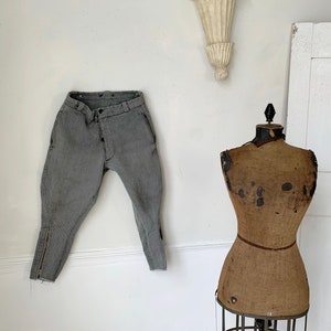 May include: A pair of gray riding breeches with a zipper detail on the legs hangs on a white wall next to a vintage brown fabric dress form.