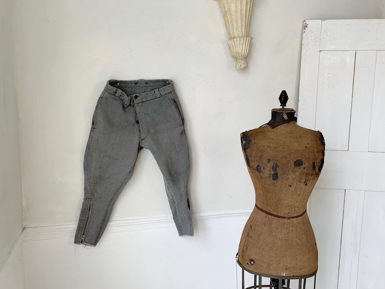 May include: A pair of gray riding breeches with a zipper detail on the legs hangs on a white wall next to a vintage brown fabric dress form.