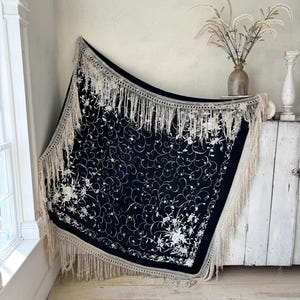 May include: A black shawl with white embroidery and fringe. The shawl is draped over a white surface and has a floral design.