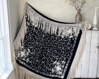 May include: A black shawl with white embroidery and fringe. The shawl is draped over a white surface and features a floral design.