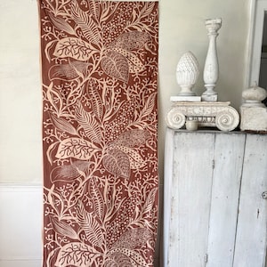 Può includere: Una tenda di tessuto marrone con un motivo floreale bianco. Il motivo presenta foglie e fiori stilizzati. La tenda è appesa a una parete accanto a un armadio bianco con una parte superiore decorativa.