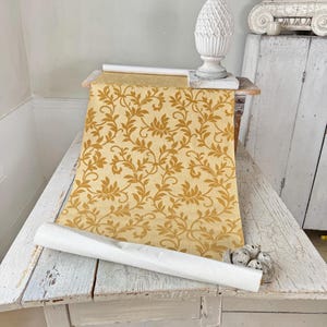 Puede incluir: Un rollo de papel pintado con un diseño floral dorado y crema, desenrollado sobre una mesa de madera blanca desgastada. También se ven una estatua de piña blanca y una pequeña escultura. Ideal para la decoración de interiores.