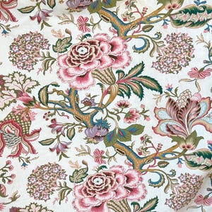 Op de afbeelding: Een witte stof met een herhalend bloemenpatroon. Het patroon bevat roze, rode, groene en blauwe bloemen met ingewikkelde details. De achtergrond is crèmekleurig.
