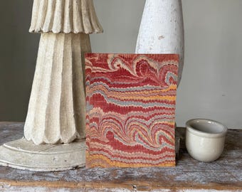 Può includere: Un taccuino rettangolare con una copertina marmorizzata nei toni del rosso, giallo e grigio. Il taccuino è esposto su una superficie di legno usurata, accanto a un vaso di ceramica bianca e una piccola ciotola bianco sporco. È presente anche una statua decorativa scolpita bianco sporco.