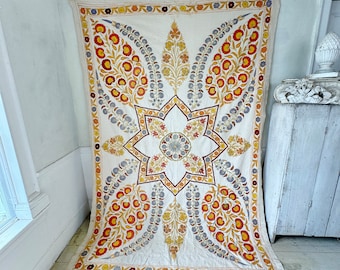 May include: A white cotton fabric wall hanging with a large star-shaped design in the center. The star is surrounded by four floral motifs in shades of orange, yellow, and blue. The fabric has a frayed edge.