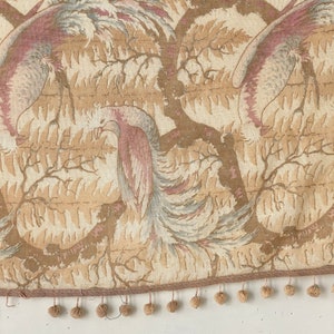 Op de afbeelding: Een beige stof met een herhalend patroon van roze en grijze vogels in een bosrijke omgeving. De stof heeft een bruine franje met kleine pompoenen.