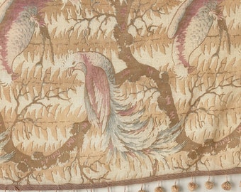May include: A beige fabric with a repeating pattern of pink and gray birds in a forest setting. The fabric has a brown fringe trim with small pom-poms.