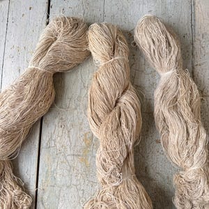 May include: Three bundles of natural-colored yarn, tightly wound and tied with twine. The yarn has a slightly textured appearance and is arranged on a weathered, light-colored wooden surface. The bundles are of varying sizes and shapes.