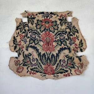 Pode incluir: Um pedaço de tecido bege com um padrão floral em vermelho, verde e azul. O design apresenta grandes flores e folhas estilizadas. As bordas do tecido estão inacabadas.