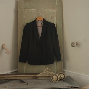 May include: A black blazer hanging on a wooden door with a light brown hanger. The blazer has a notched lapel and a single button closure. The door is painted a light green color.