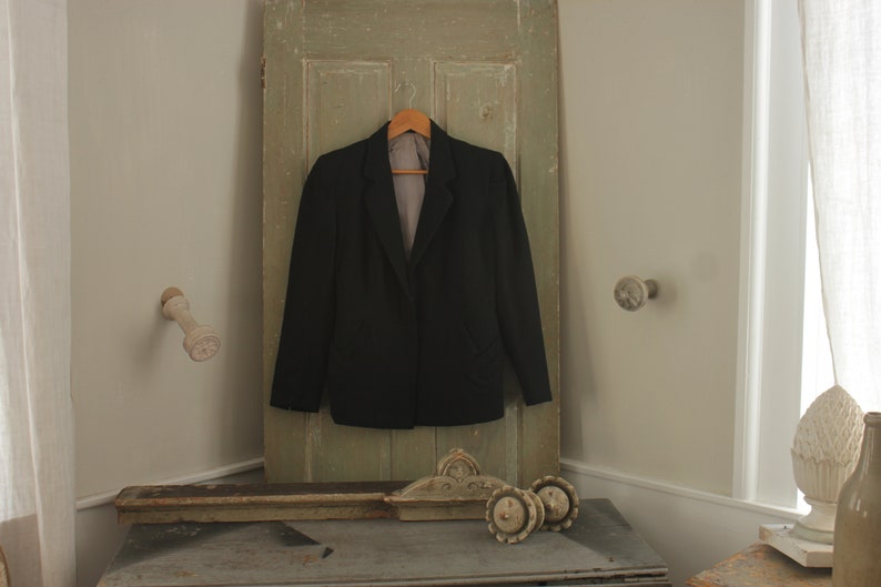 May include: A black blazer hanging on a wooden door with a light brown hanger. The blazer has a notched lapel and a single button closure. The door is painted a light green color.