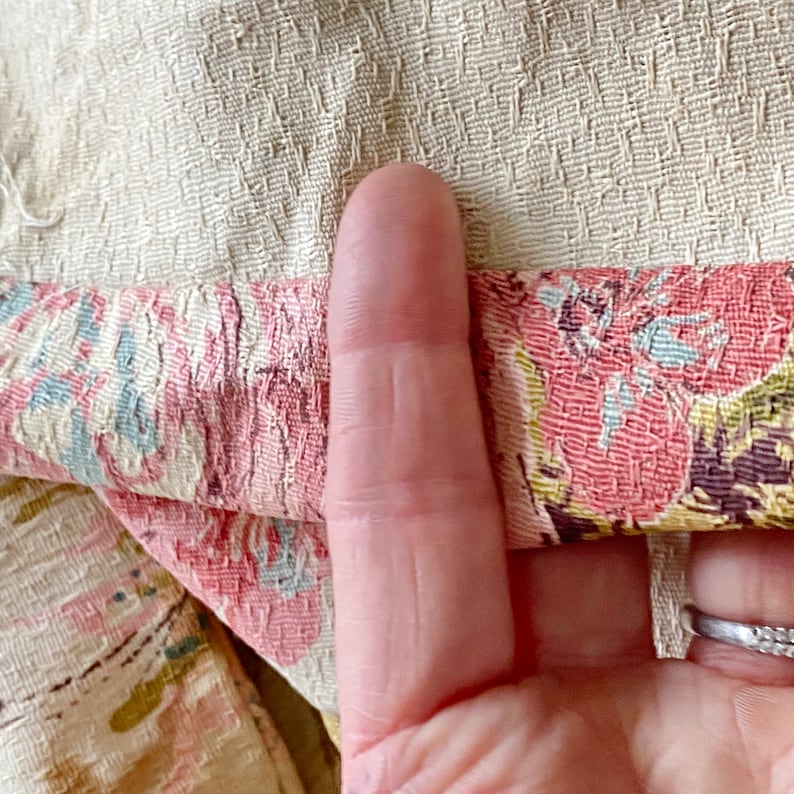 May include: Close-up of a cream-colored fabric with a floral pattern in shades of pink, blue, and green. The fabric has a textured surface and is folded over.