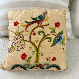 May include: A beige pillow with a colorful embroidered design of a bird perched on a tree branch with flowers and leaves.