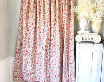 May include: A floral patterned curtain in shades of pink, blue, and cream. The curtain features a repeating pattern of flowers and leaves, with a border along the edges. The curtain is hanging in front of a wall.
