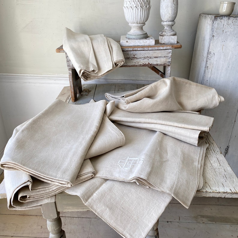 May include: A stack of off-white linen tablecloths, some with embroidered initials, on a white wooden table.