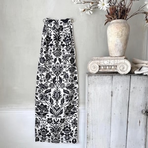Op de afbeelding: Een lange gordijn met een zwart-wit bloemenpatroon, hangend aan een muur. Het ontwerp is symmetrisch, met gedetailleerde bloemen en bladeren. Een vaas met bloemen en een decoratieve zuil staan op de achtergrond.