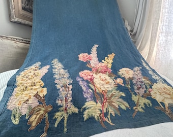 May include: A long, blue textile with a floral design. The embroidered flowers include pink, yellow, and purple blooms with green leaves. The textile is draped over a white surface.