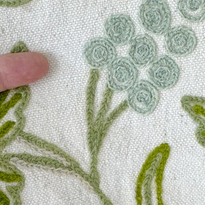 May include: Close-up of a white fabric with green embroidered floral designs. The design features a cluster of small, round, light green flowers with stems and leaves.