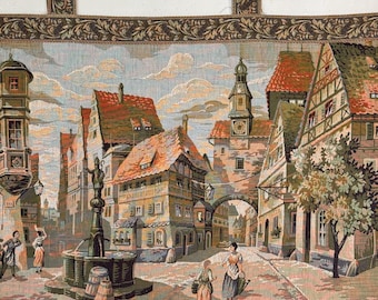 Könnte beinhalten: Eine Tapisserie, die einen europäischen Marktplatz mit bunten Gebäuden, einem Brunnen und Spaziergängern darstellt. Die Tapisserie hat einen braunen und grünen Rand mit einem floralen Muster.
