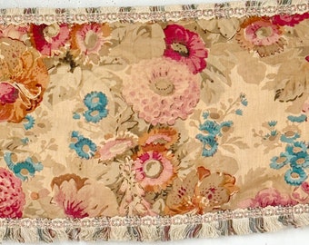 May include: A beige fabric trim with a floral pattern featuring pink, orange, and blue flowers. The trim has a fringe edge.