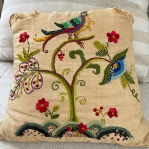 May include: A beige pillow with a hand-stitched embroidery design featuring a stylized tree with a bird, flowers, and leaves. The embroidery is done in shades of green, red, blue, purple, and yellow.