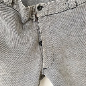 May include: Close-up of the waistband and fly of a pair of gray corduroy pants. The pants have a button fly and a single button at the top of the fly.