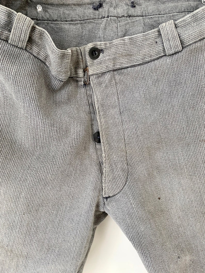May include: Close-up of the waistband and fly of a pair of gray corduroy pants. The pants have a button fly and a single button at the top of the fly.
