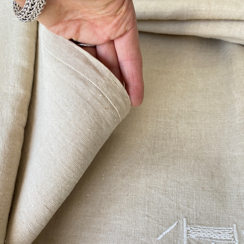 May include: A close-up of a natural linen tablecloth with a subtle texture. The fabric is folded over, revealing the underside of the cloth.