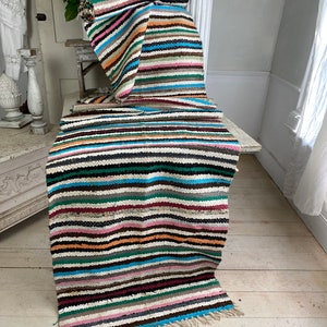 Op de afbeelding: Een kleurrijk gestreept tapijt met een franje aan de onderkant. Het tapijt is gemaakt van geweven stof en heeft een verscheidenheid aan kleuren, waaronder bruin, zwart, wit, blauw, groen, roze en oranje.