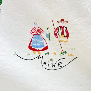 May include: White linen tablecloth with embroidered figures of a woman and a man in traditional clothing. The woman is wearing a red and blue dress and the man is wearing a brown hat and a red vest. The word "MAINE" is embroidered in black below the figures.