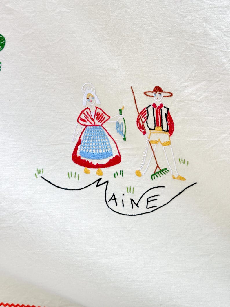 May include: White linen tablecloth with embroidered figures of a woman and a man in traditional clothing. The woman is wearing a red and blue dress and the man is wearing a brown hat and a red vest. The word "MAINE" is embroidered in black below the figures.